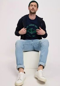 Granatowa bluza z zamkiem błyskawicznym na ciemnej koszulce z grafika zielonej góry, jasnoniebieskie dżinsy i białe sneakersy. Model siedzi na białym sześcianie.