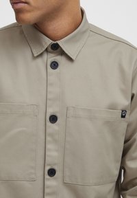 Beige shirt featuring a button-down collar, two chest pockets, and black buttons. Notable brand tag on the left pocket. Smooth fabric texture.