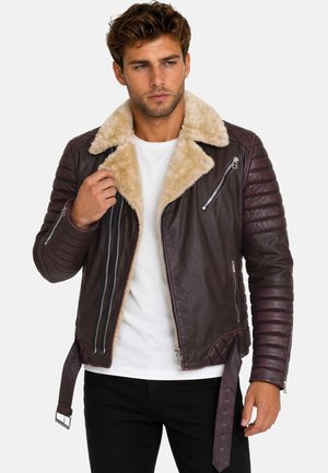 Hombre con chaqueta de cuero marrón con cuello de piel beige sobre camisa blanca y pantalones negros, ajustando el cuello de la chaqueta con la mano izquierda.