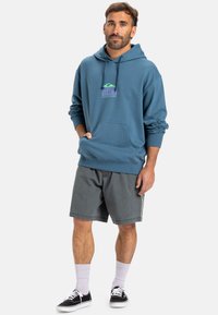 Sweat à capuche bleu avec une poche avant et un logo graphique. Porté avec un short gris, des chaussettes blanches et des baskets noires. Tissu doux, coupe décontractée.