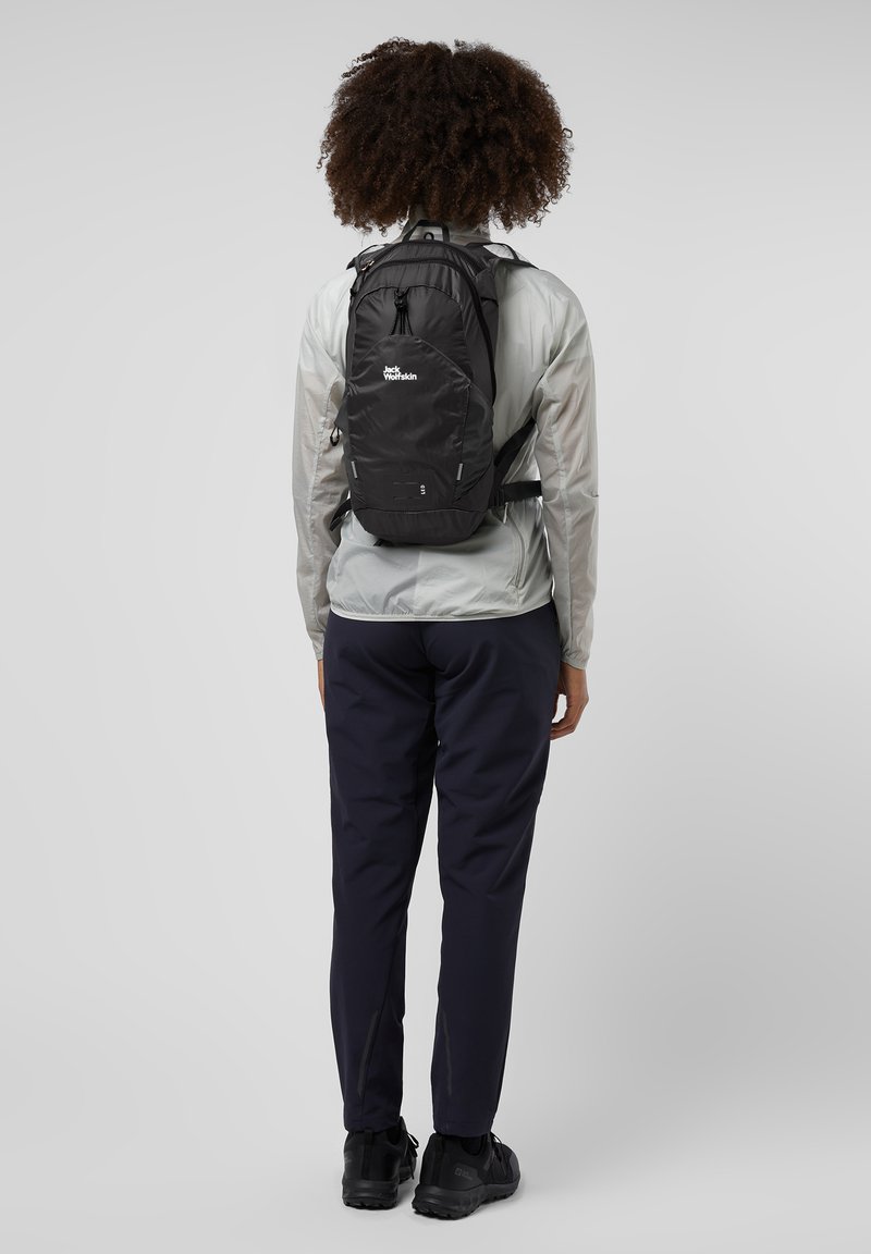 Schwarzer Rucksack mit einem eleganten Design, verstellbaren Riemen und Logo. Trägt man über eine leichte Jacke und dunkle Hosen, vor einem grauen Hintergrund.