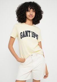 Sárga pamut póló "GANT 1949" felirattal tengerészkék színben. Világos bézs rövidnadrággal párosítva, amelyen gomb és két zseb található.