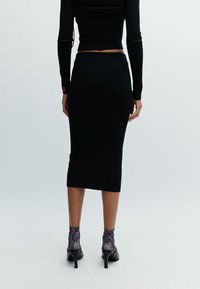 Femme se tenant de dos, portant un haut court noir à manches longues, une jupe midi noire, des chaussettes courtes à motifs et des chaussures à talons noirs.