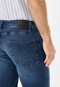 Jean en denim bleu foncé ajusté avec une patch en cuir noir sur la taille, comportant deux poches arrière et des détails de couture verticale.