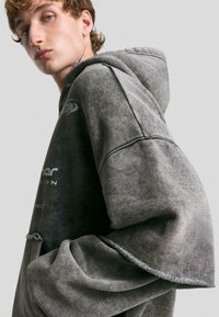 Gray oversized hoodie with a smooth texture. Features a drawstring hood, logo accents, and a relaxed fit. Visible stitching detail.