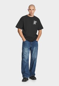 Czarna bawełniana koszulka z białym emblematem na lewej piersi, w połączeniu z luźnymi niebieskimi dżinsami i czarnymi trampkami.