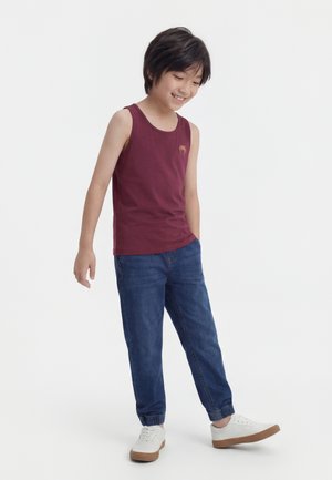 Garçon souriant portant un débardeur bordeaux, un jean jogger bleu et des baskets blanches, debout avec un pied légèrement levé.