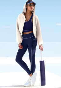 Jūras spēka sporta apģērbs, kas sastāv no īsās blūzes, augsta vidukļa legginsiem ar baltām šuvēm, apvienots ar krēmkrāsas kapuci un baltām sporta kedām, tur rokās kriketa nūju.