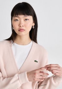 Cardigã rosa claro com decote em V, fecho de botão e logotipo bordado verde. Usado sobre uma t-shirt branca, apresenta mangas largas.