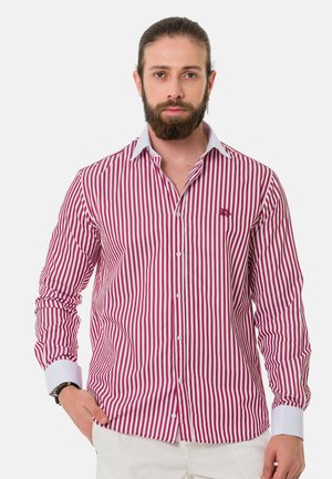 Man met baard en teruggebonden haar, draagt een rood-wit verticaal gestreept overhemd met witte kraag en manchetten, staand met één hand in de zak.