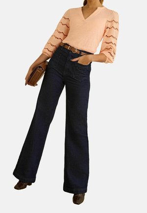 Femme portant un chemisier pêche à col en V avec des motifs ondulés en dentelle sur les manches trois-quarts, un jean large foncé, une ceinture marron, des chaussures marron et tenant une pochette marron.