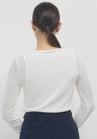 Weißes Langarm-Strickoberteil mit strukturiertem Muster und weitem Rundhalsausschnitt, kombiniert mit dunklen, taillierten, hochgeschnittenen Hosen mit Knopfdetails.