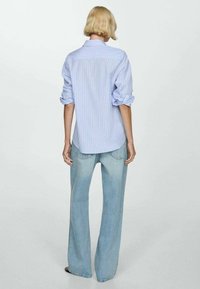 Chemise à rayures bleu clair avec manches retroussées, vue de dos ; associée à un pantalon large en jean clair, mettant en valeur une silhouette décontractée.