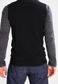 Svart ärmlös tröja med hög krage, kombinerad med långa grå ärmar. Slätt tyg, ribbat fåll och sidokragde accent.