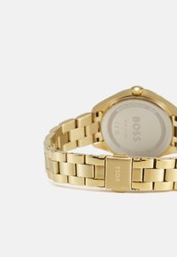 Montre analogique en or avec un bracelet en maillons métalliques. Présente un cadran rond, une texture lisse et un fermoir avec le nom de la marque gravé.