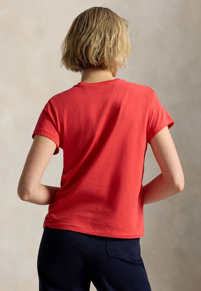 Rövid szőke hajú személy vörös rövid ujjú pólót és sötét nadrágot visel, kezei a csípőjén, háttal egy egyszínű háttér előtt áll.