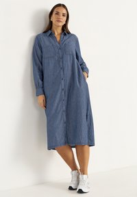 Denim dress with long sleeves, chest pockets, button-up front design, a loose silhouette, and a length that reaches mid-calf. Paired with white sneakers.