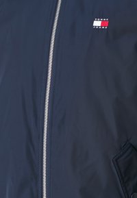 Tmavě modrá bunda na zip s hladkou texturou, s emblémem v barvách červené, bílé a modré na levé straně hrudi a boční kapsou.