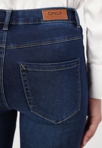 Un jean en denim bleu foncé présente une patch en cuir avec l'étiquette "ONLY" à la taille, ainsi qu'une poche arrière ornée de coutures courbes.