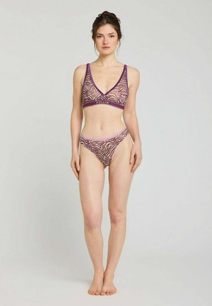 Femme debout pieds nus portant un ensemble soutien-gorge et culotte imprimé zèbre violet et rose sur un fond gris clair uni.