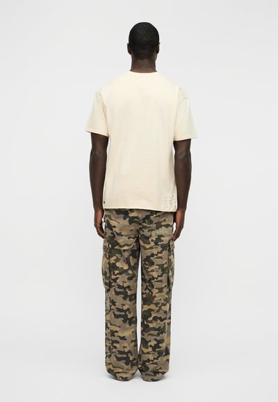Φωτεινό μπεζ βαμβακερό μπλουζάκι με κοντά μανίκια, σε συνδυασμό με χαλαρά παντελόνια καμουφλάζ τύπου cargo με πολλαπλές τσέπες και ρυθμιζόμενο τελείωμα με κορδόνι.