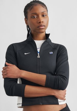 Noor isik, kellel on punutud juuksed ja keda ehtib must, tõmblukuga Nike logoga jakk, käed rüpes, selja taga helehall ühevärviline taust.