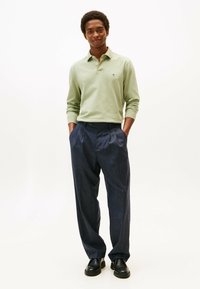 Camisa polo de manga larga verde claro con un pequeño logo, combinada con pantalones de traje oscuros con pliegues y zapatos negros.