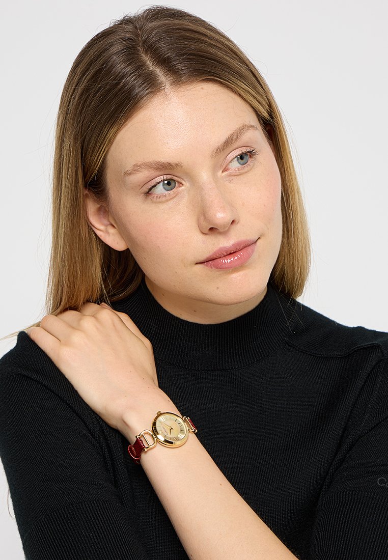 Montre en or avec un cadran rond et un bracelet en cuir rouge, présentant un cadran minimaliste et des accents métalliques. La montre est portée sur un poignet.