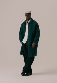 Grønn rutete ullkåpe med stor krage, beige strikket skjorte, svarte bukser og glansfulle sorte sko, toppet med en lysfarget cap.