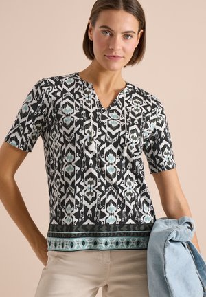 Frau trägt ein schwarz-weiß-türkis gemustertes V-Ausschnitt-Shirt mit kurzen Ärmeln und hält in ihrer linken Hand eine hellblaue Jeansjacke.