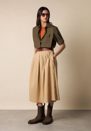 Vrouw met een oversized olijfgroen overhemd over een oranje top, beige midi-rok met knopen, bruine riem, donkere laarzen en zonnebril, staand tegen een effen achtergrond.