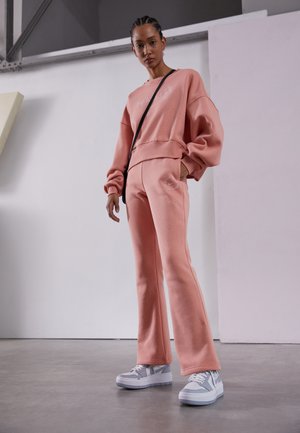 Sweatshirt rosa claro com texto bordado, combinado com calças de fato correspondentes que apresentam um logótipo. Ténis brancos com pormenores em cinza. Textura suave.