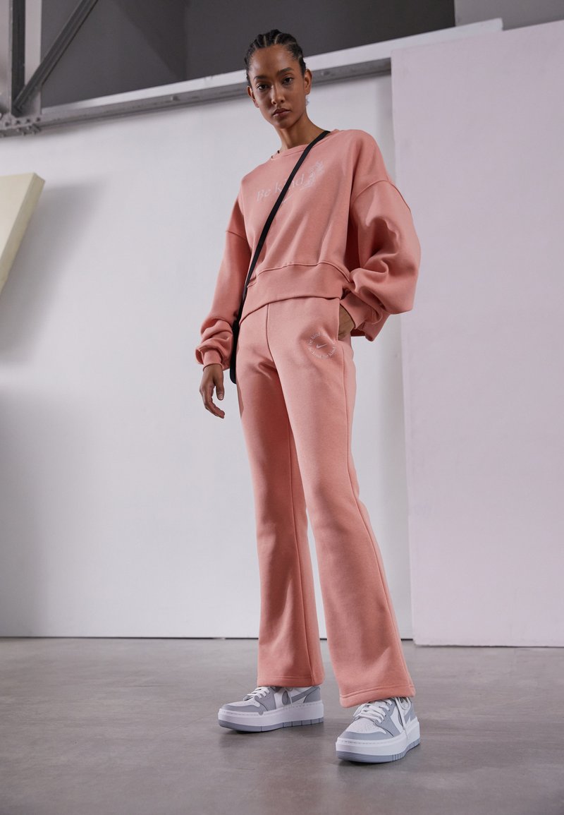 Sweatshirt rosa claro com texto bordado, combinado com calças de fato correspondentes que apresentam um logótipo. Ténis brancos com pormenores em cinza. Textura suave.