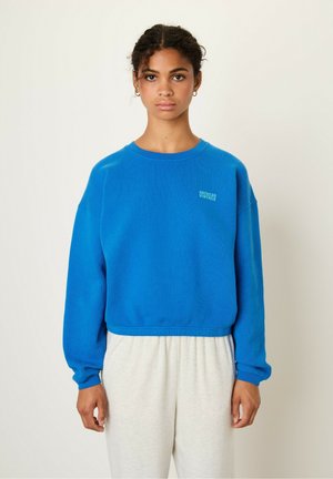 Blauer, kurz geschnittener Sweatshirt aus Fleece-Material, mit Rundhalsausschnitt und gerippten Bündchen. Kleines Logo auf der linken Brust. Kombiniert mit hellgrauen Hosen.