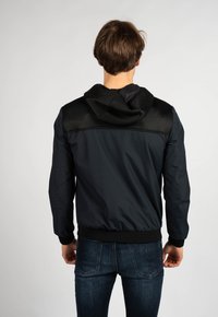 Giacca blu scuro con cappuccio a rete nera testurizzata e dettagli sulle spalle. Presenta un design aderente e polsini elasticizzati, realizzata in materiale leggero.