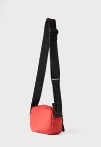 Czerwona nylonowa torba na ramię z zamkiem błyskawicznym, kwadratowym kształtem i regulowanym czarnym paskiem z tłoczonym logo.