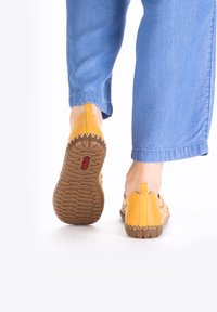 Gelbe Leder-Slipper mit einem geflochtenen Design, Gummisohle und einer kleinen Fersenschlaufe. Jeans sind am Knöchel teilweise sichtbar.