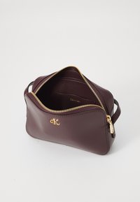 Bordo kožna torba s patentnim zatvaračem i zlatnim detaljima; ima bočnu ručku i glatku teksturu s logotipom unutra.