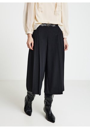 Femme portant un pantalon culotte noir ample avec une ceinture noire, un chemisier crème à manches longues et des bottes en cuir noires à bout pointu.