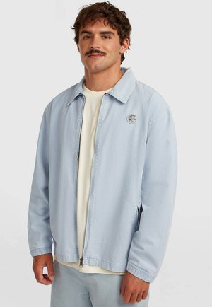 Svetlomodrá bunda z tkaniny zmesi bavlny, s stojatým golierom, predným zipsom a nášivkou s logom na ľavej strane hrudníka.