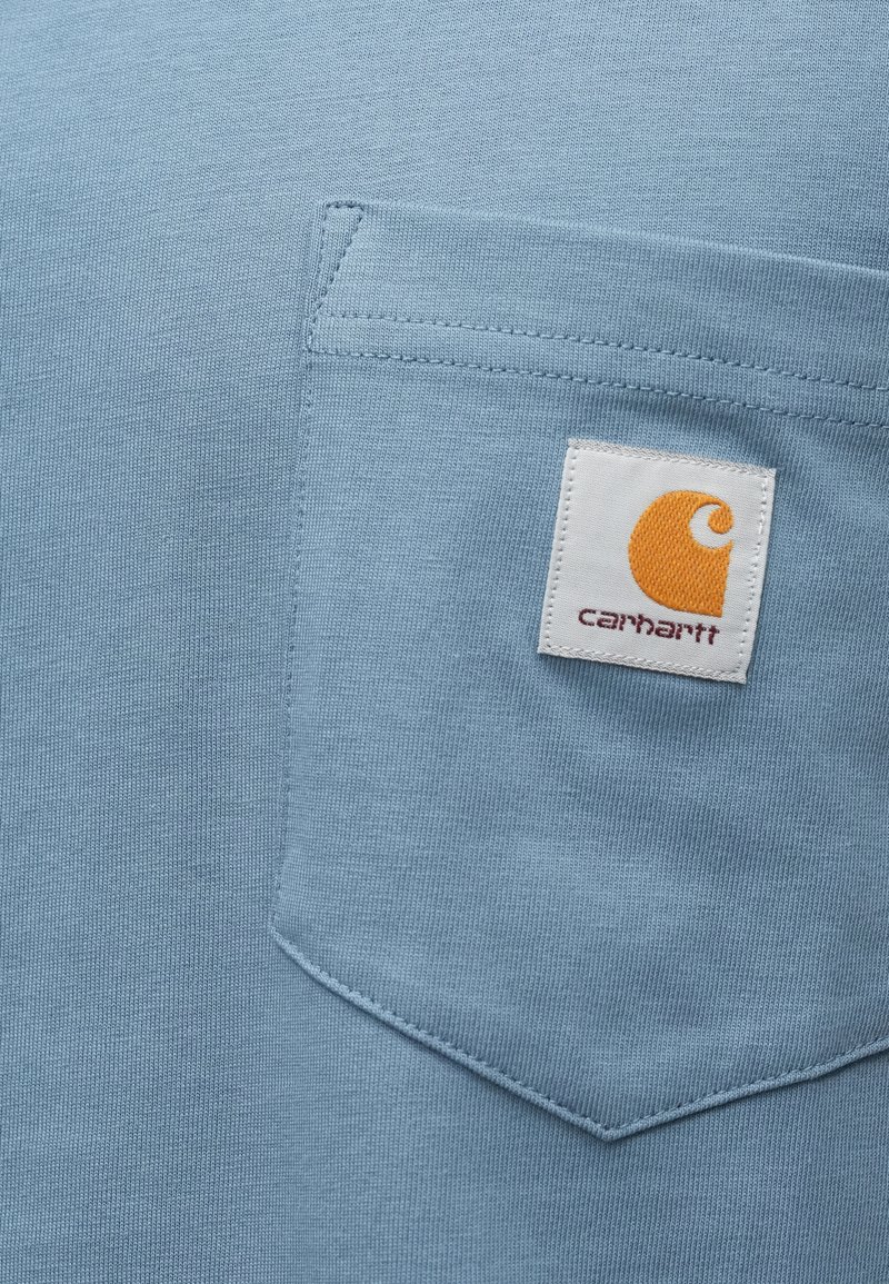 Lys blå T-shirt med en brystlomme med hvid stikning og et firkantet orange logo-patch med et stiliseret bogstav "C."
