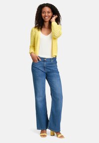 Cardigan tricoté jaune sur un top blanc, associé à un jean bleu taille haute et large, et des sandales à talons jaunes. Pose souriante, main sur la hanche.