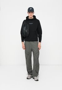 Sort hættetrøje med logo, grå cargo bukser med store lommer, sort crossbody taske, grå sneakers og en sort kasket.
