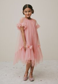 Helles rosa Tüllkleid mit Rüschensleeves und Schleifen an der Taille. Getragen mit glitzernden pinken Schuhen. Einfacher Hintergrund mit subtilen Dekorationen.