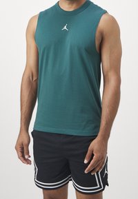 Top sin mangas en color verde azulado con cuello redondo y logo blanco de Jordan, combinado con pantalones cortos de malla negros que presentan franjas blancas y detalles de Jordan.