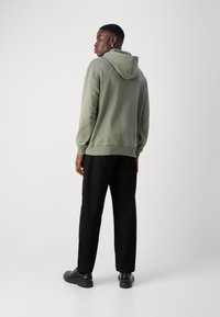 Haut à capuche vert olive avec cordon de serrage, poignets et ourlet côtelés ; associé à un pantalon noir et des chaussures noires, vu de derrière.