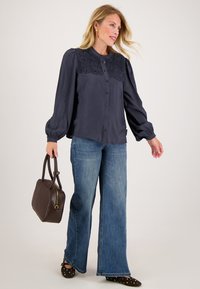 Marineblouse met een kant yoke, knoopsluiting aan de voorkant, puffed mouwen, gecombineerd met wijde blauwe jeans en een bruin handtas. Detail van de schoenen met stippen is zichtbaar.