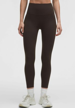 Vrouw draagt zwarte leggings met hoge taille, witte sokken en witte sportschoenen, staand tegen een effen lichte achtergrond.