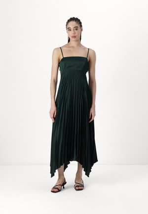 Abito verde con pieghe, spalline sottili, tessuto fluido, orlo asimmetrico e una texture liscia, abbinato a sandali neri con cinturino.