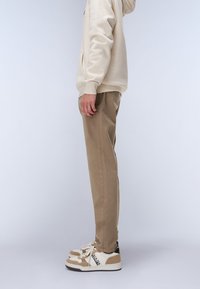 Beige hoodie med en framficka, parat med skräddarsydda khakibyxor och vita sneakers med beige och svarta detaljer.